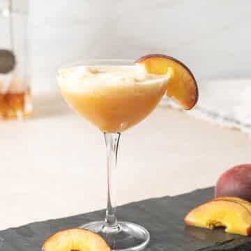 A coupe glass filled with a peach-colored cocktail, garnished with a peach slice on the rim, sits on a slate surface with additional peach slices nearby.