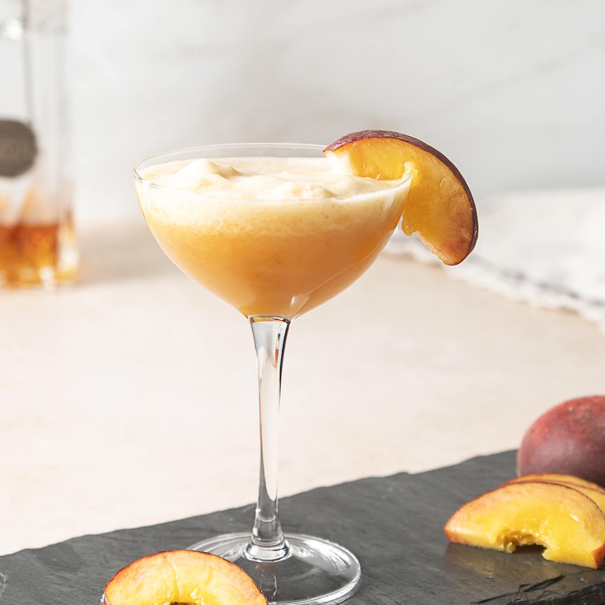 A coupe glass filled with a peach-colored cocktail, garnished with a peach slice on the rim, sits on a slate surface with additional peach slices nearby.