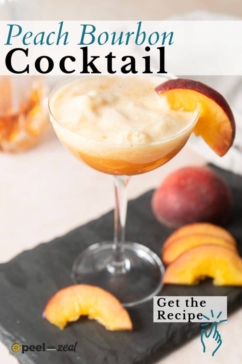 A peach bourbon cocktail in a coupe glass, garnished with a peach slice, sits on a slate board with peach wedges and a whole peach in the background.