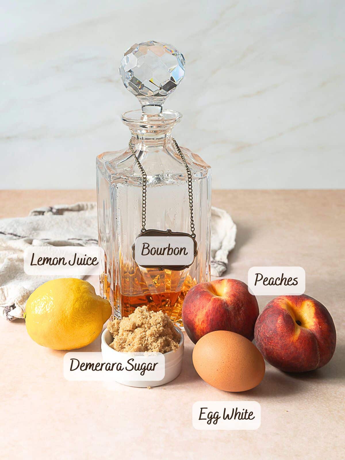 A decanter of bourbon, a lemon, fresh peaches, a bowl of demerara sugar, and an egg are arranged on a light countertop. Each item is labeled with text.