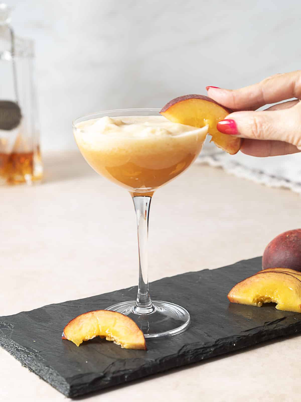 A hand garnishes a frothy peach-colored cocktail in a coupe glass with a fresh peach slice; more peach slices are on the slate serving board.
