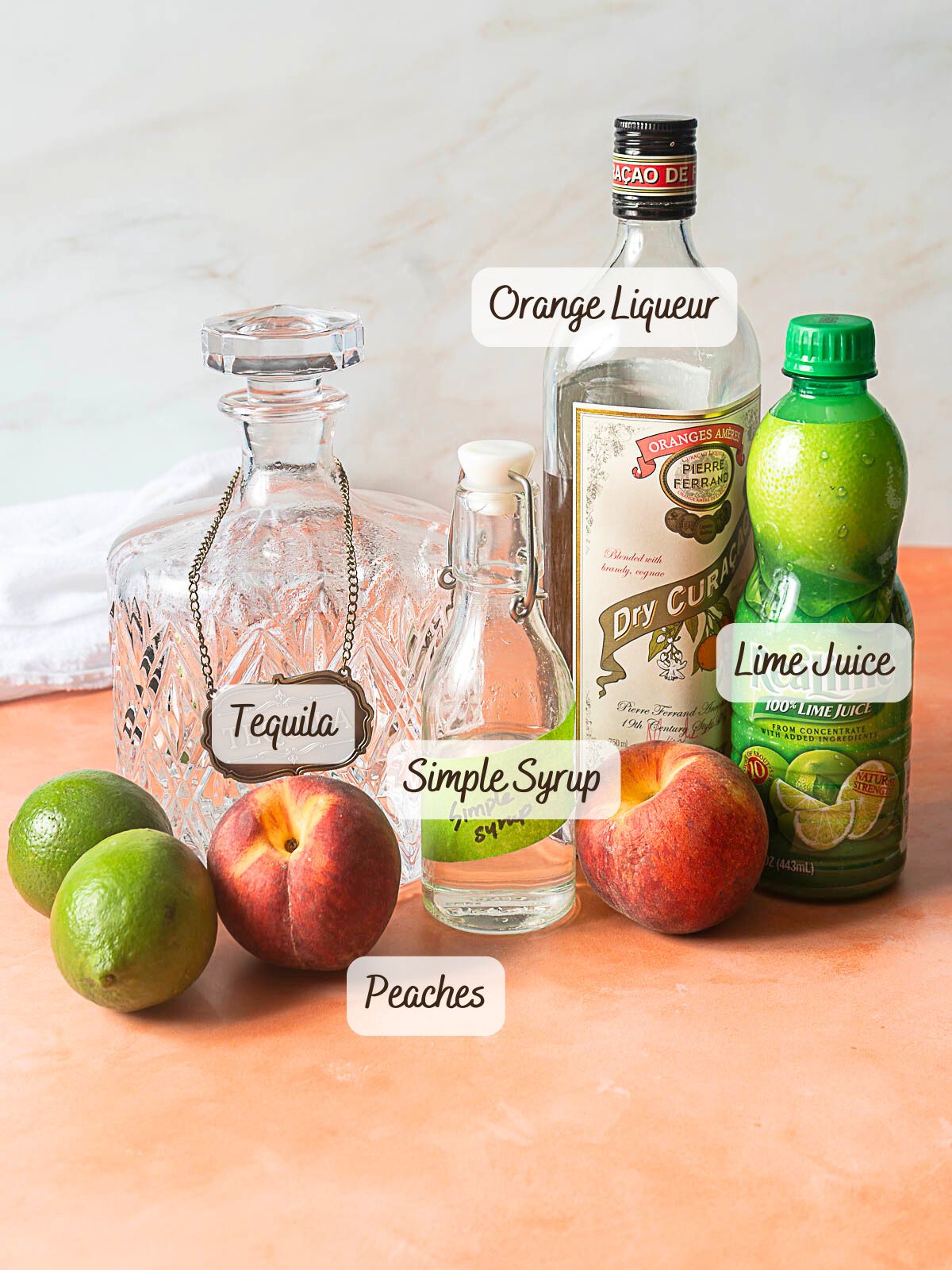 A bottle of orange liqueur, a decanter labeled tequila, simple syrup, lime juice, two limes, and two peaches are arranged on a counter.