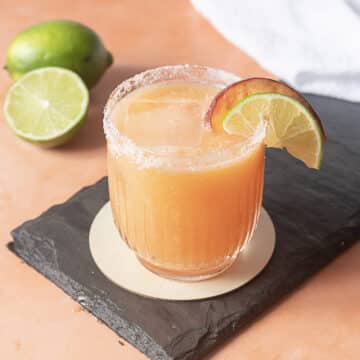 A glass of orange-colored cocktail with a salted rim, garnished with a lime and peach slice, sits on a slate coaster. A halved lime is in the background.
