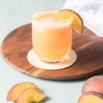 A glass of peach-colored drink with ice and a peach slice garnish sits on a wooden tray, with extra peach slices nearby.