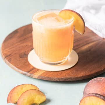 A glass of peach-colored drink with ice and a peach slice garnish sits on a wooden tray, with extra peach slices nearby.