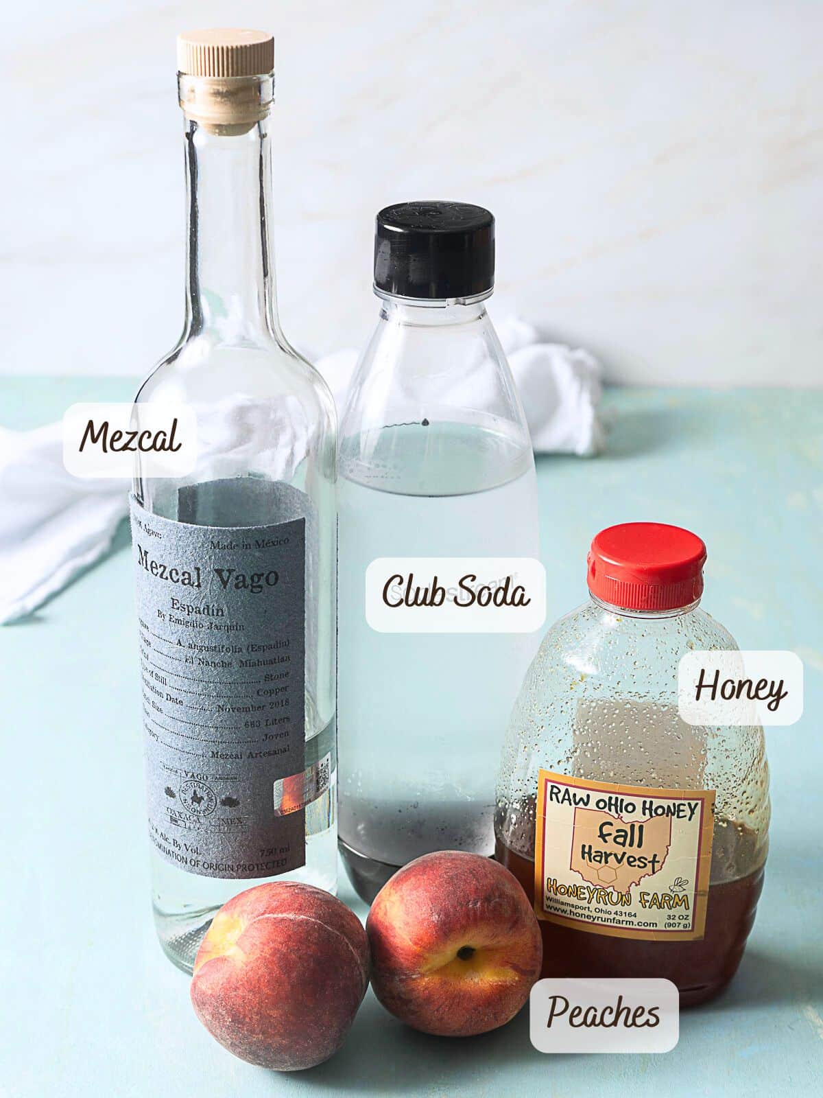 A bottle of mezcal, a bottle of club soda, a jar of honey, and two peaches are arranged on a light blue surface. Each item is labeled.