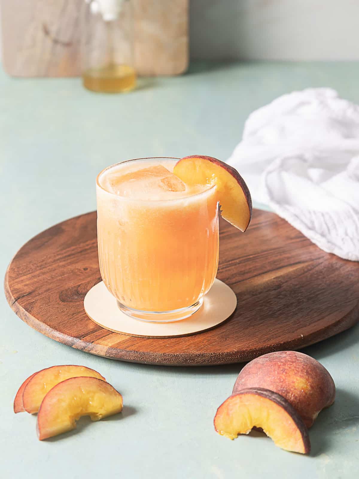 A glass of peach-colored beverage with ice and a peach slice garnish sits on a round wooden tray, with peach slices and a whole peach nearby.