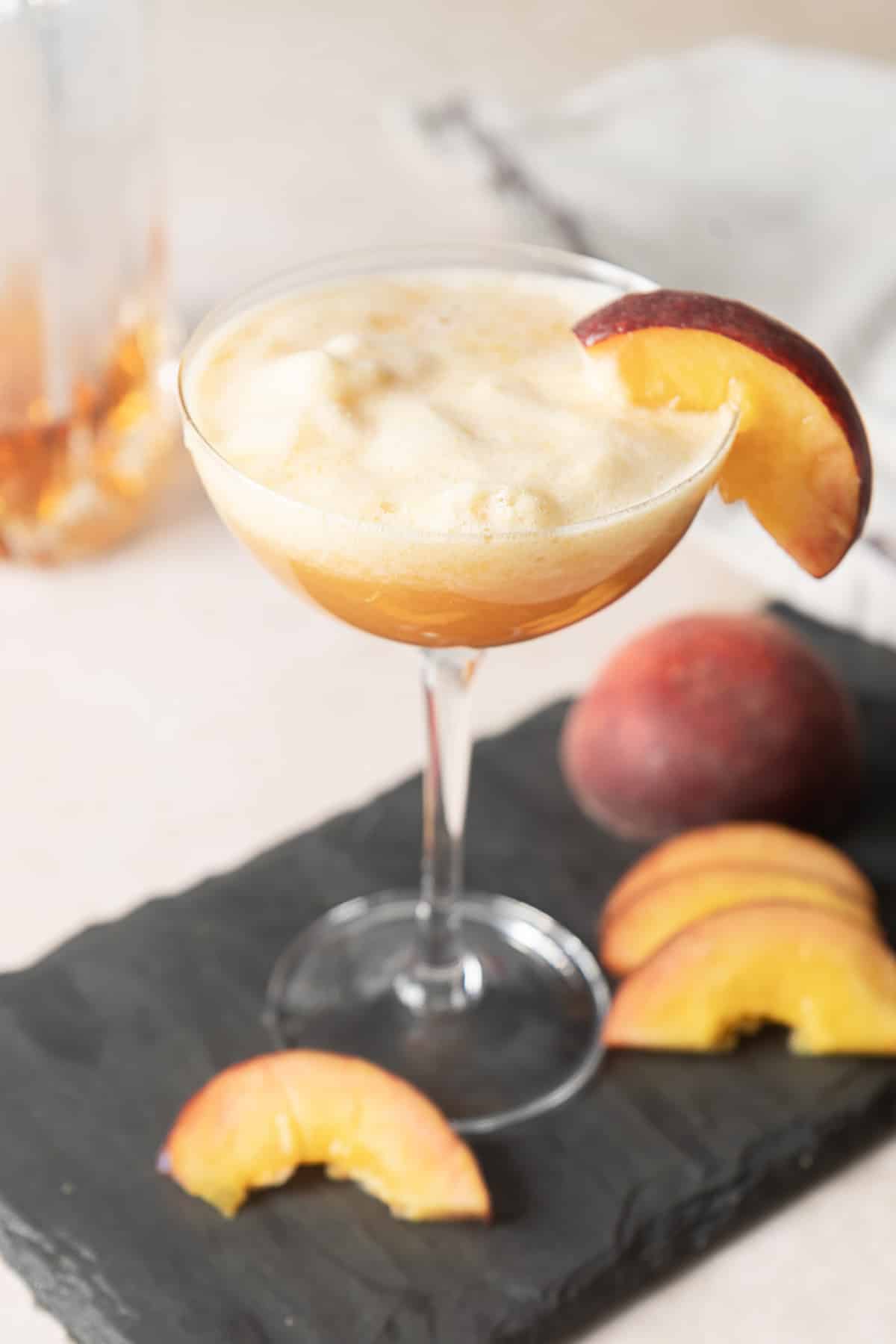 A coupe glass filled with a frothy peach cocktail, garnished with a peach slice, sits on a slate serving board with additional peach slices and a whole peach nearby.
