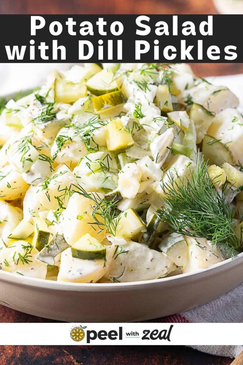 A bowl of creamy potato salad with dill pickles and fresh dill is topped with a tangy dressing, served on a wooden surface.