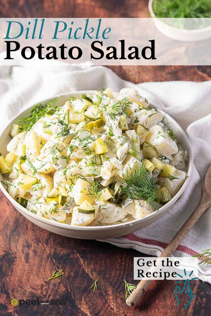 A bowl of creamy potato salad with dill pickles, garnished with fresh dill, sits on a wooden table with a wooden spoon and napkin beside it.