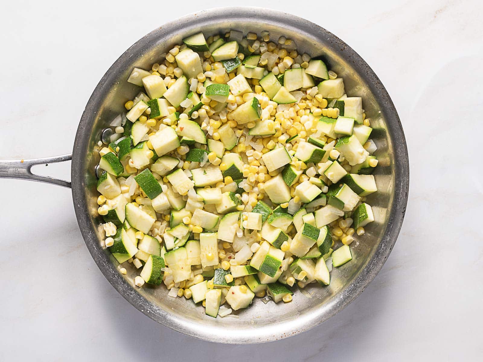 A stainless steel pan containing sautéed zucchini and corn pieces on a white surface.