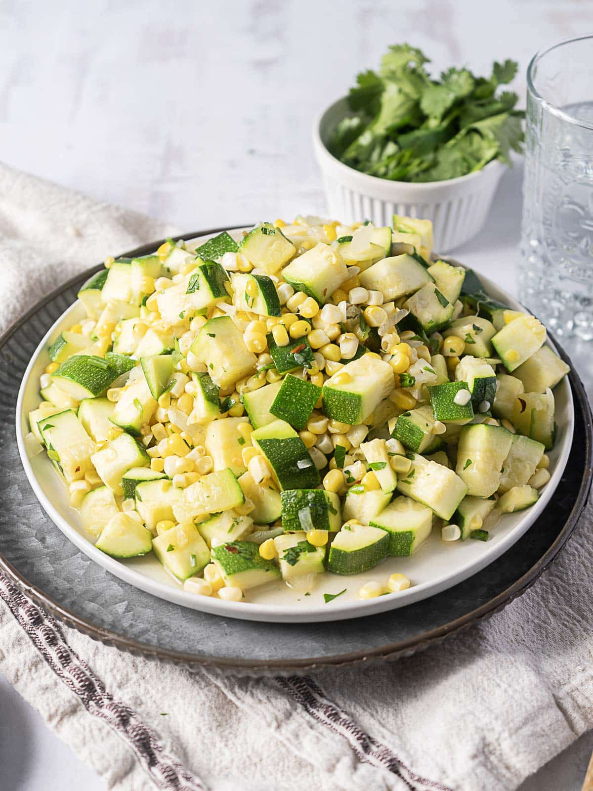 A plate of diced zucchini and corn salad is placed on a white cloth napkin, with a glass of water and a small bowl of fresh cilantro in the background.