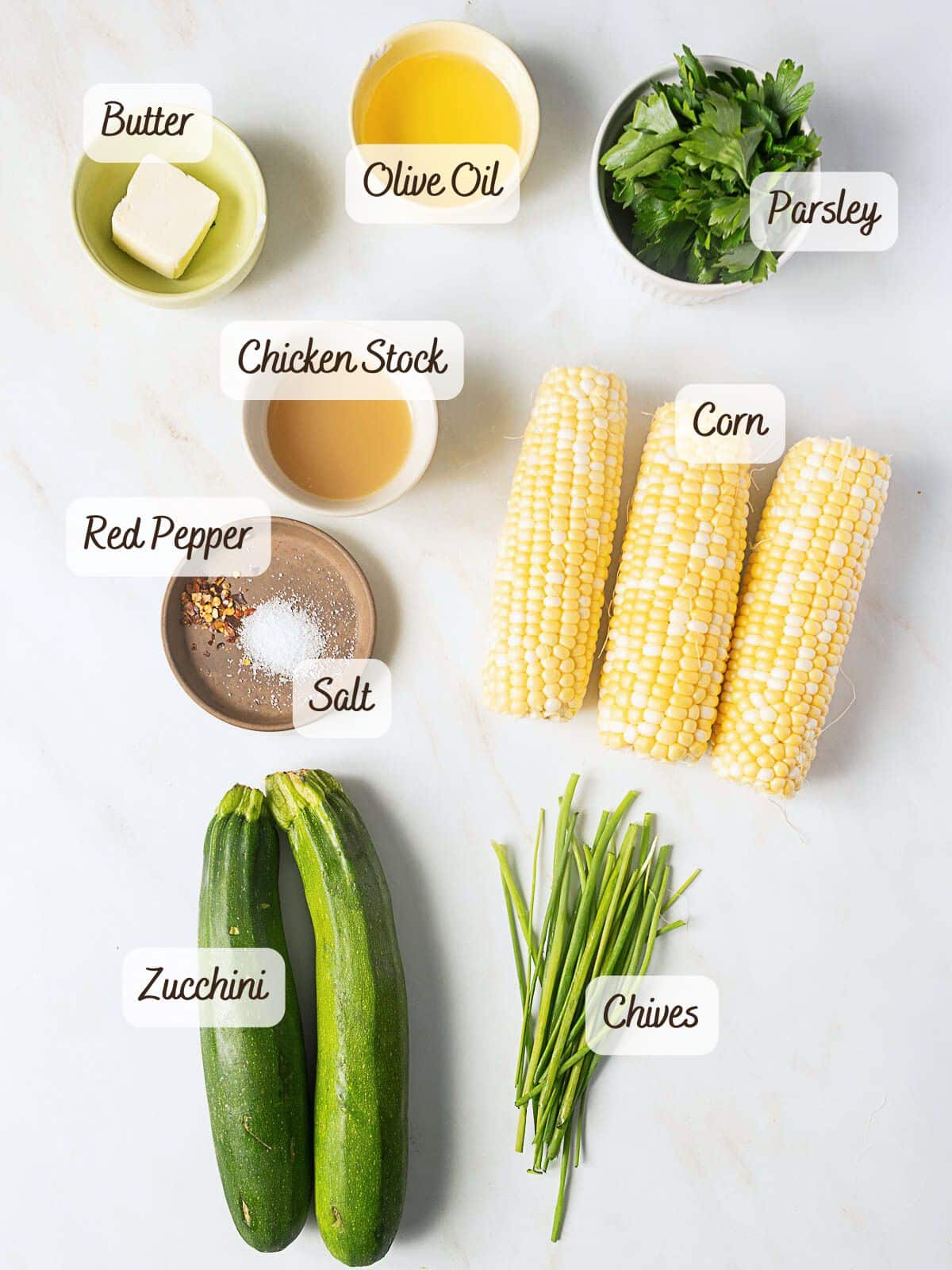 Ingredients including butter, olive oil, parsley, chicken stock, corn, red pepper, salt, zucchini, and chives are arranged on a white surface, each labeled with text.