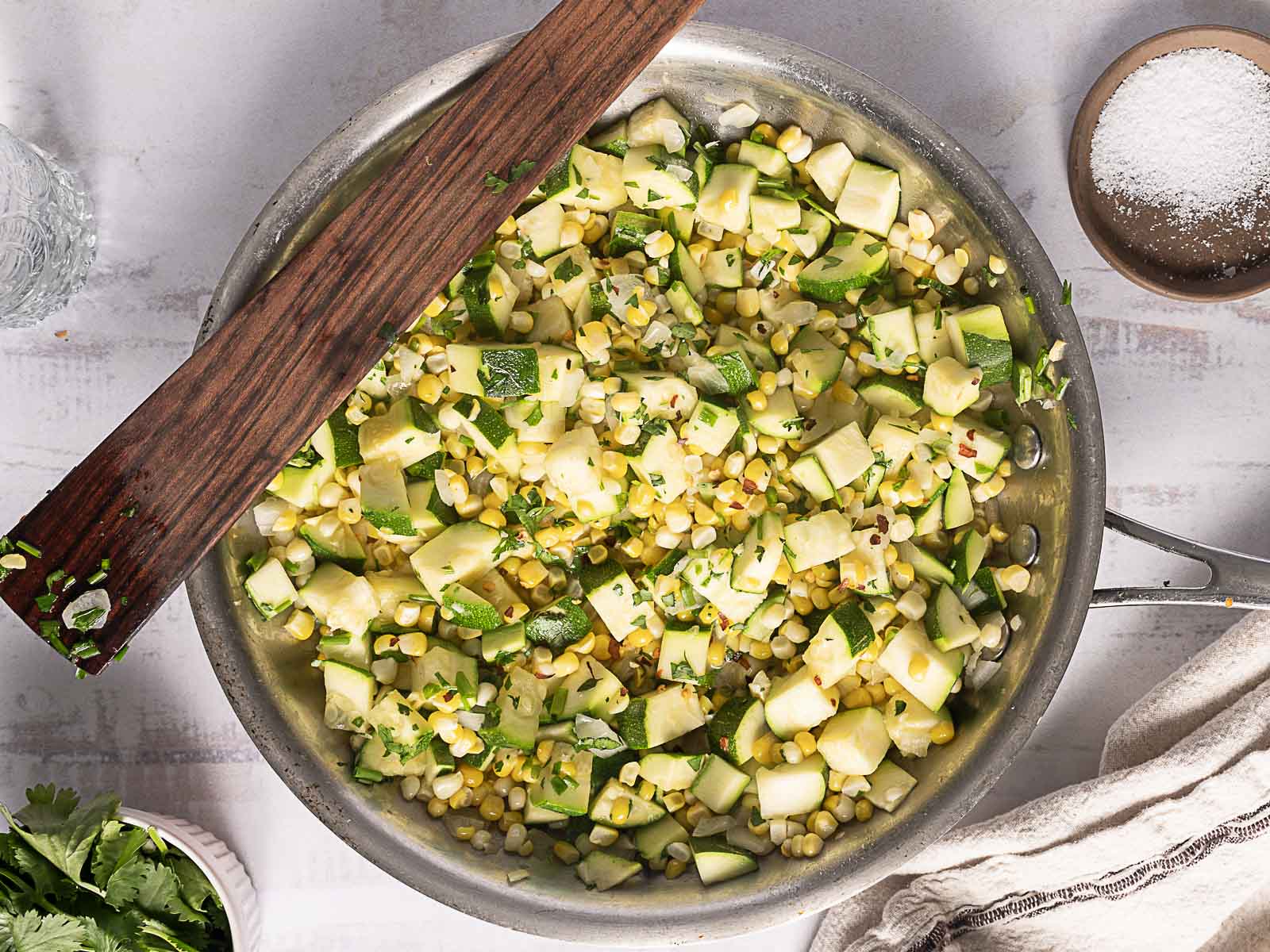 A skillet filled with sautéed corn, zucchini, and herbs, with a wooden spatula resting on top, photographed from above.