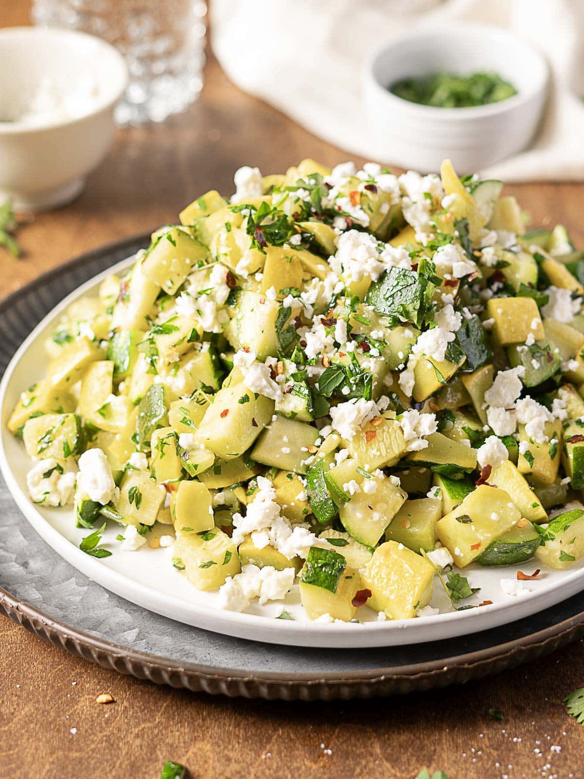 A plate of chopped zucchini salad topped with crumbled feta cheese, fresh herbs, and red pepper flakes, set on a table with small bowls and a glass of water in the background.