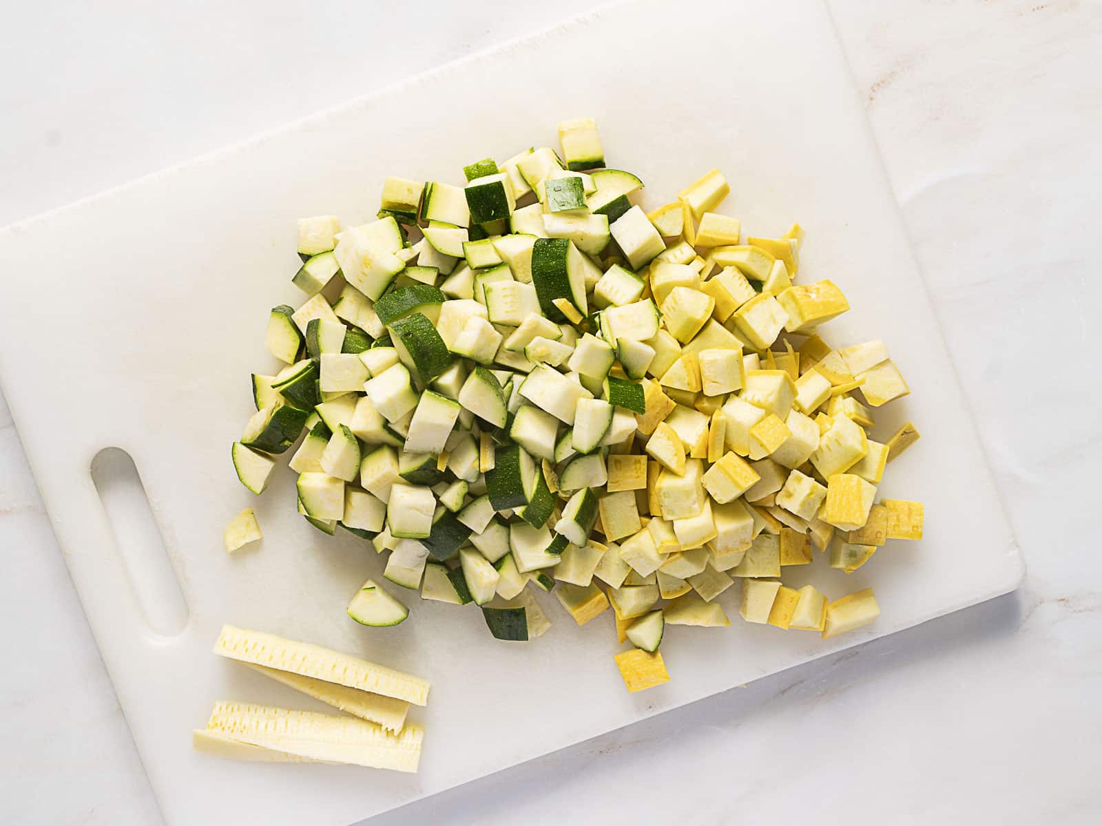 Diced green zucchini and yellow squash on a white cutting board with a few vegetable strips beside the pile.