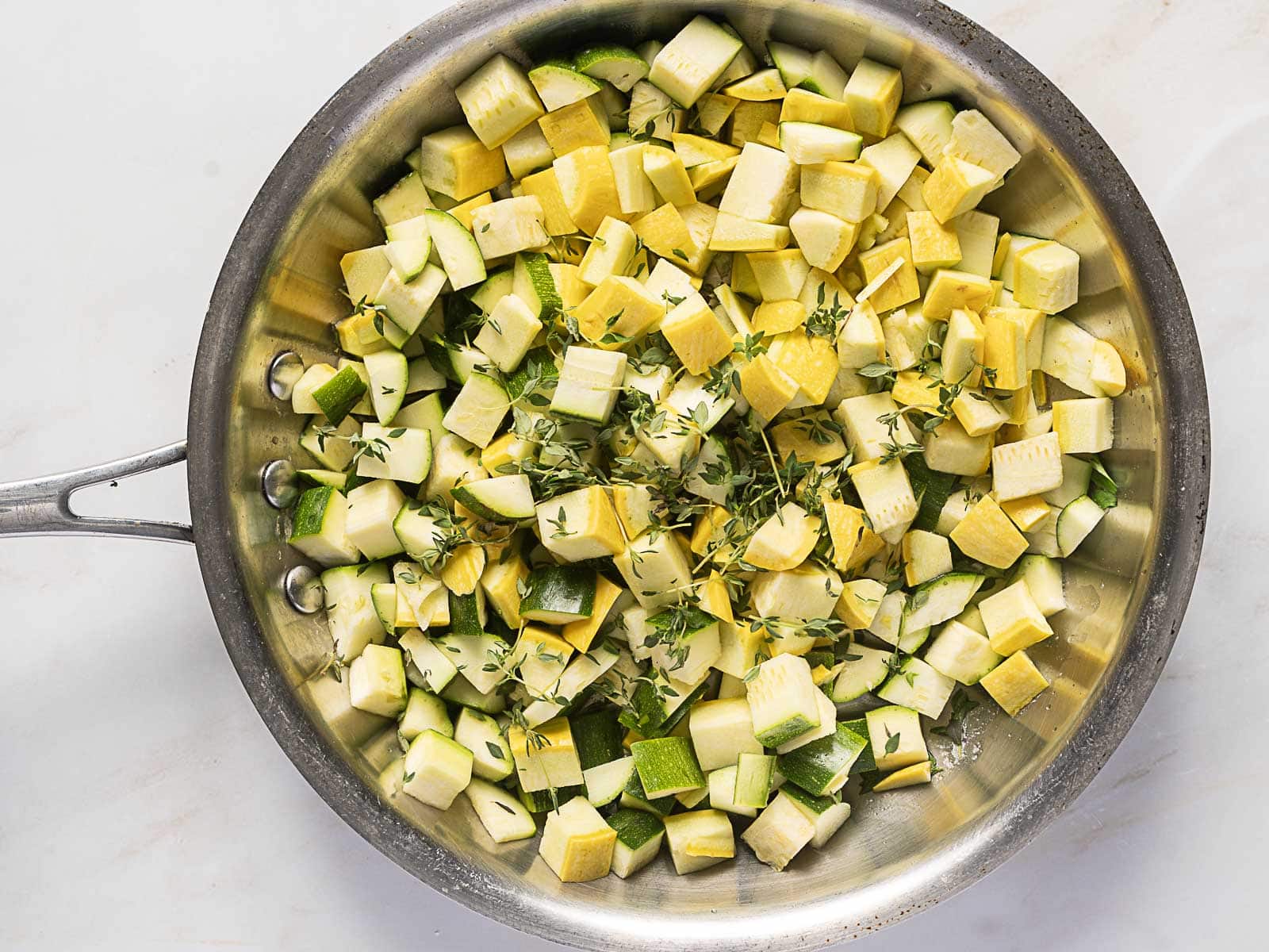Diced yellow squash and zucchini with sprigs of thyme in a stainless steel skillet, ready to be cooked.
