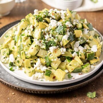 A plate of chopped zucchini salad mixed with herbs and topped with crumbled feta cheese on a brown table.