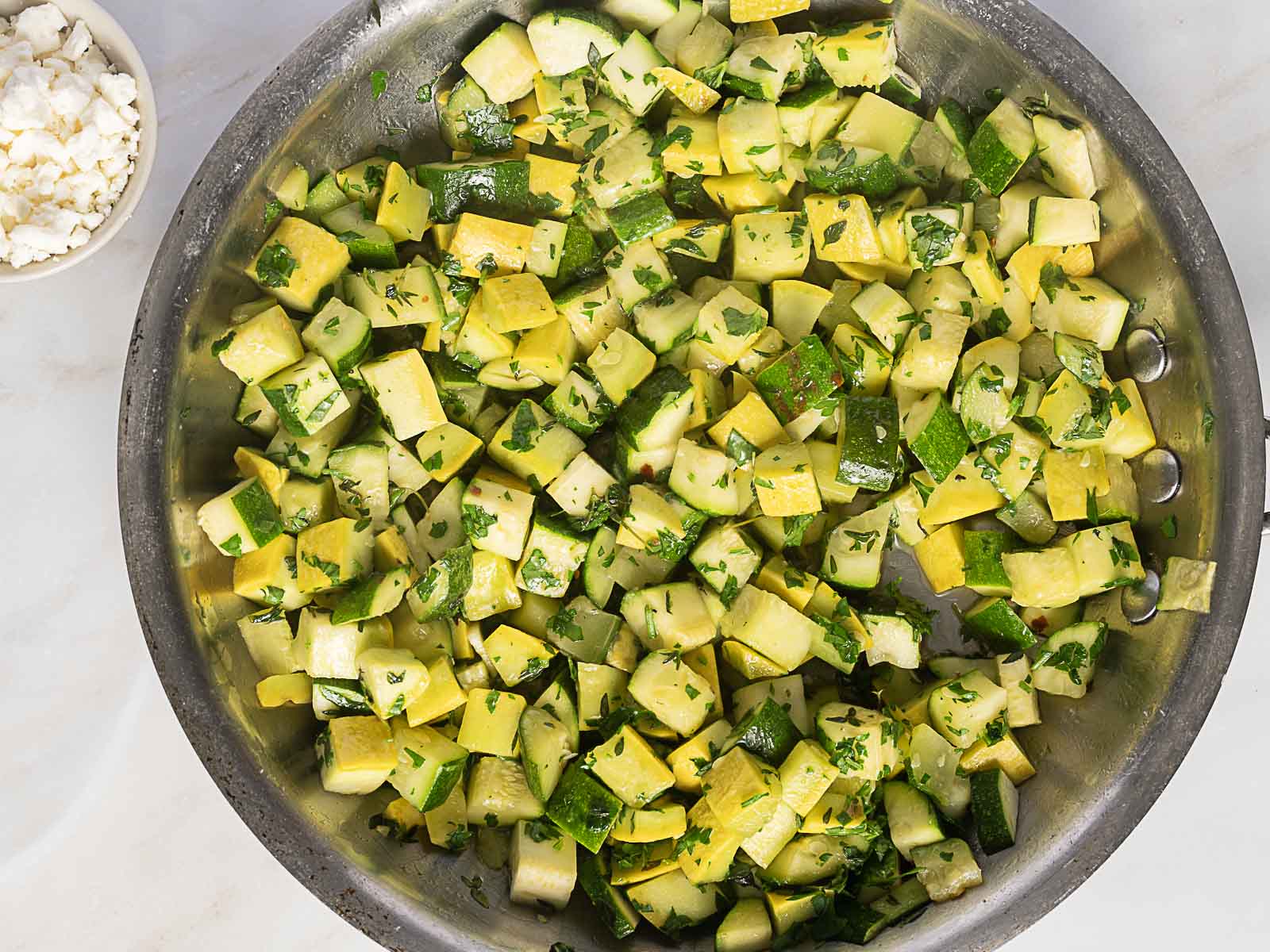 A stainless steel pan filled with sautéed diced yellow squash and zucchini, garnished with chopped herbs. A small bowl of crumbled cheese sits nearby.