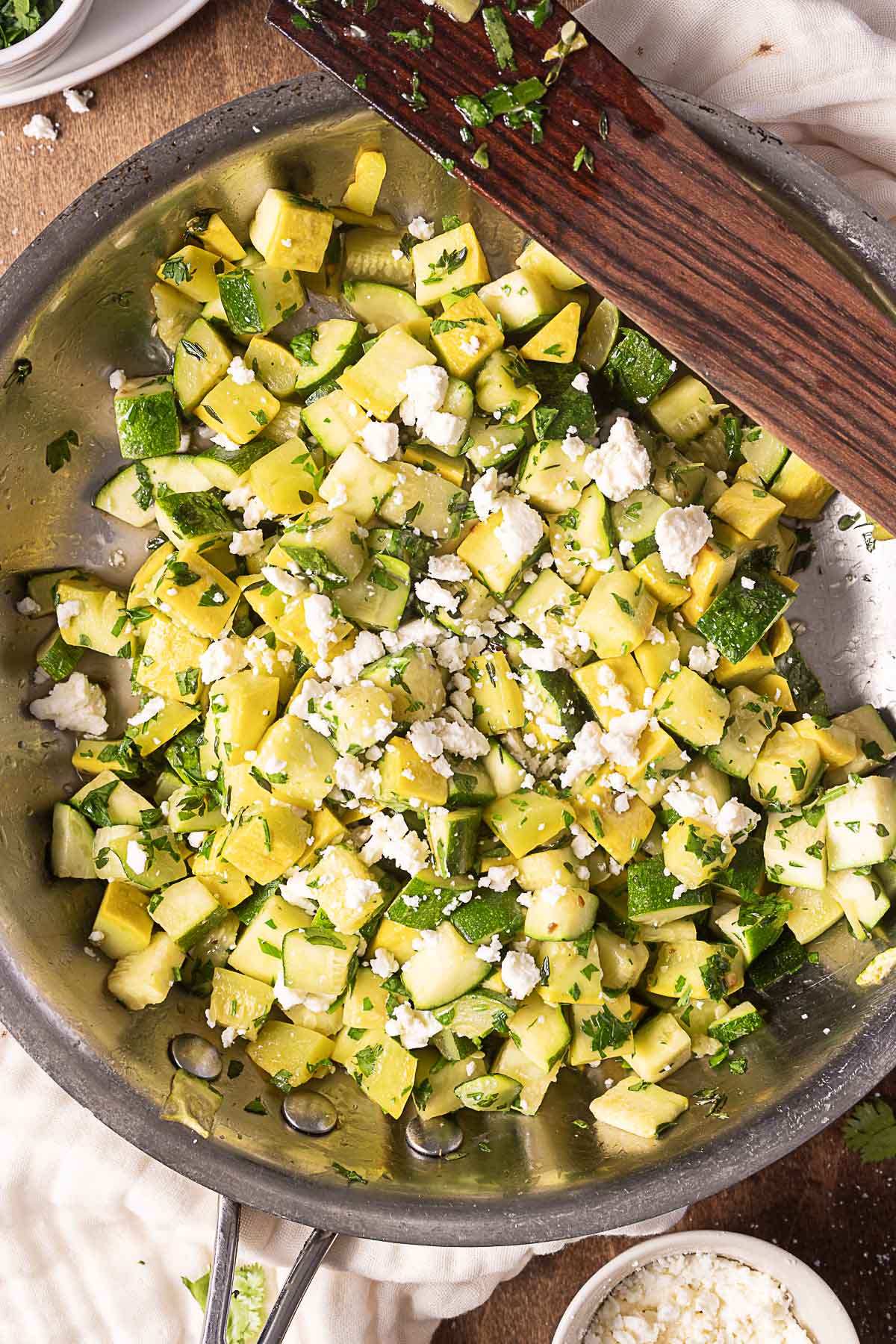 A skillet filled with sautéed diced yellow and green zucchini, topped with crumbled feta cheese and chopped parsley.