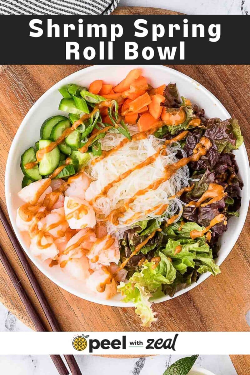 A bowl with shrimp, rice noodles, lettuce, cucumber, carrots, and scallions, topped with sauce, on a wooden surface with chopsticks beside it.