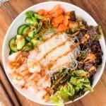 A bowl with shrimp, rice noodles, lettuce, cucumber, carrots, green onions, and peanut sauce, arranged neatly on a wooden table with chopsticks beside it.