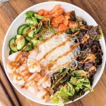 A bowl with shrimp, rice noodles, lettuce, cucumber, carrots, green onions, and peanut sauce, arranged neatly on a wooden table with chopsticks beside it.