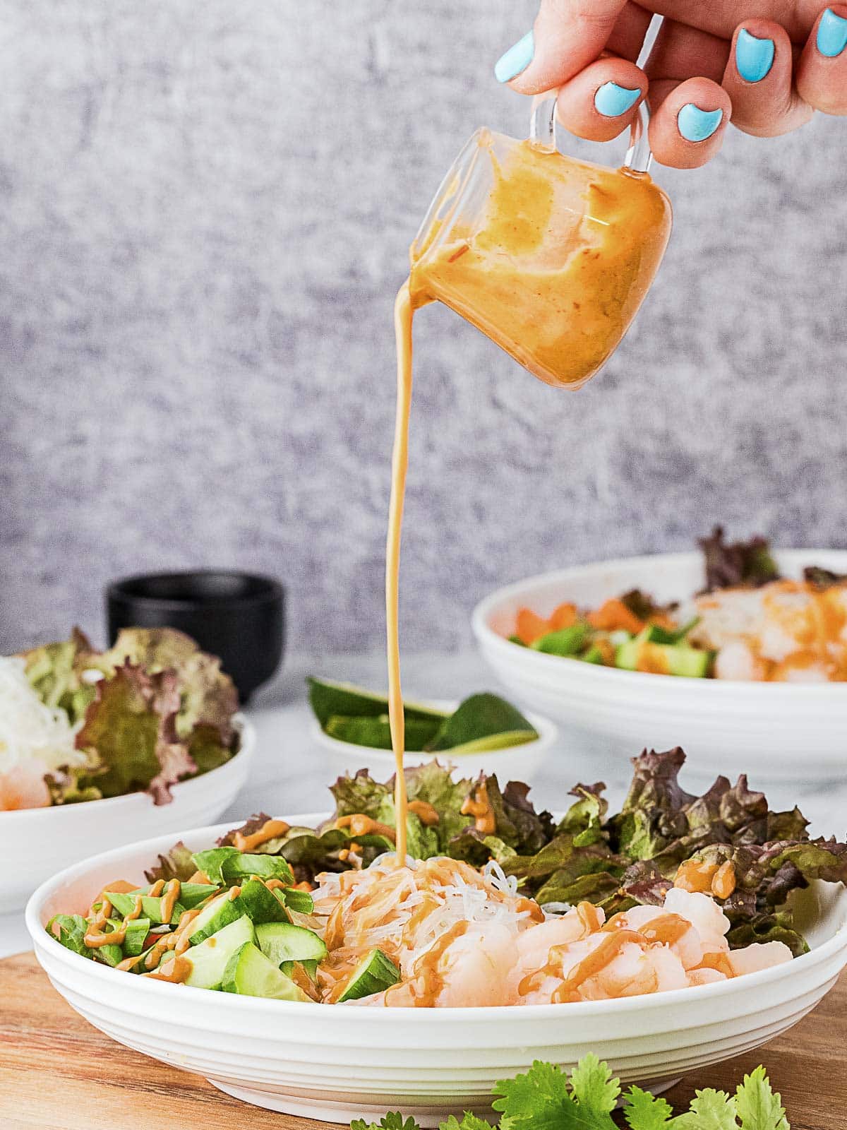 A hand pours creamy peanut sauce from a small cup onto a bowl of noodle salad with lettuce, cucumber, and chopped peanuts.