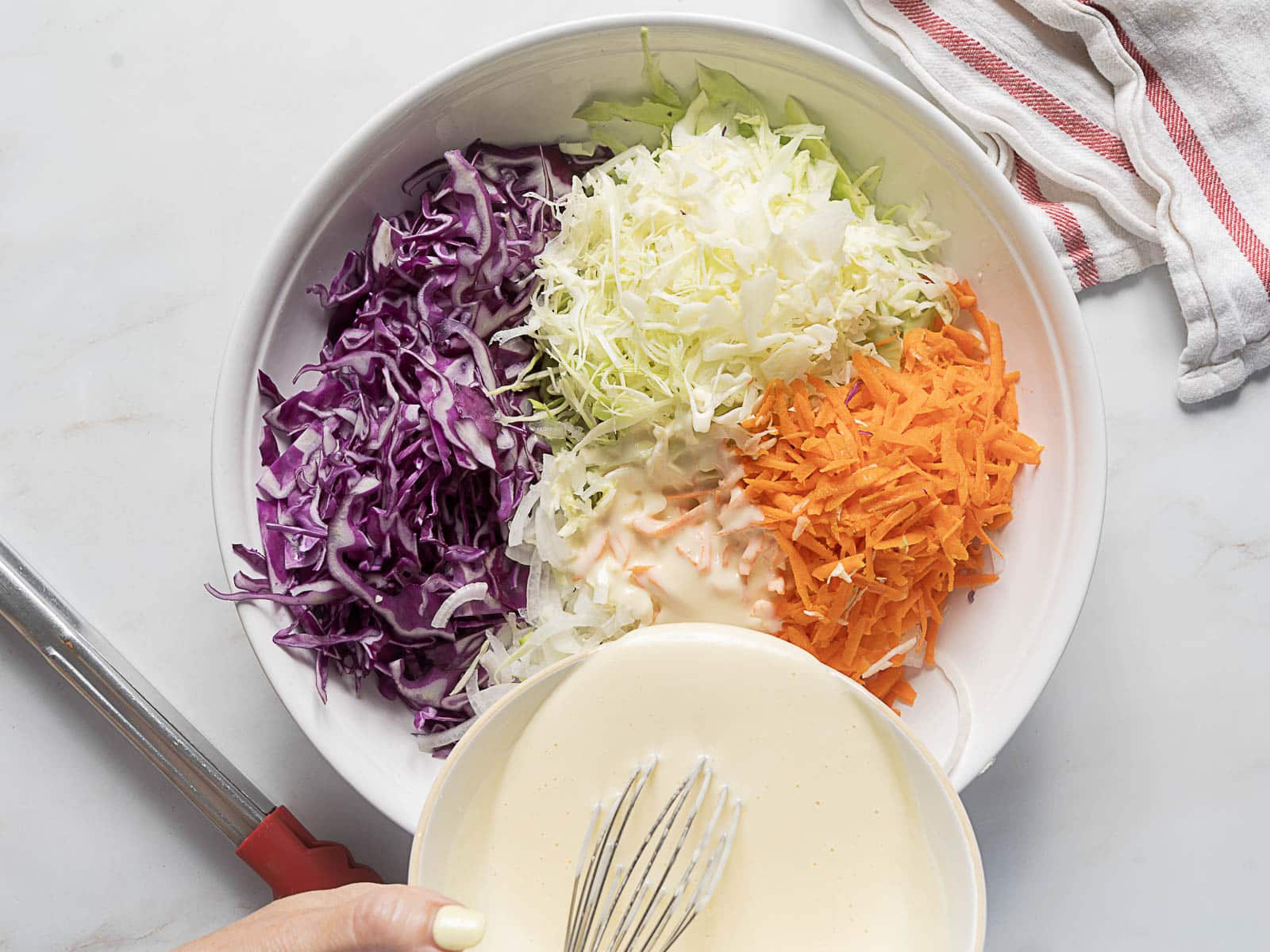 A bowl with shredded red cabbage, green cabbage, and carrots, with dressing being poured in, next to a whisk and a kitchen towel.