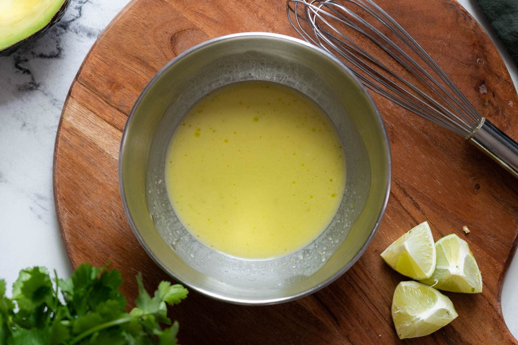 A metal bowl with yellow salad dressing, next to a whisk, lime wedges, and cilantro on a wooden board.
