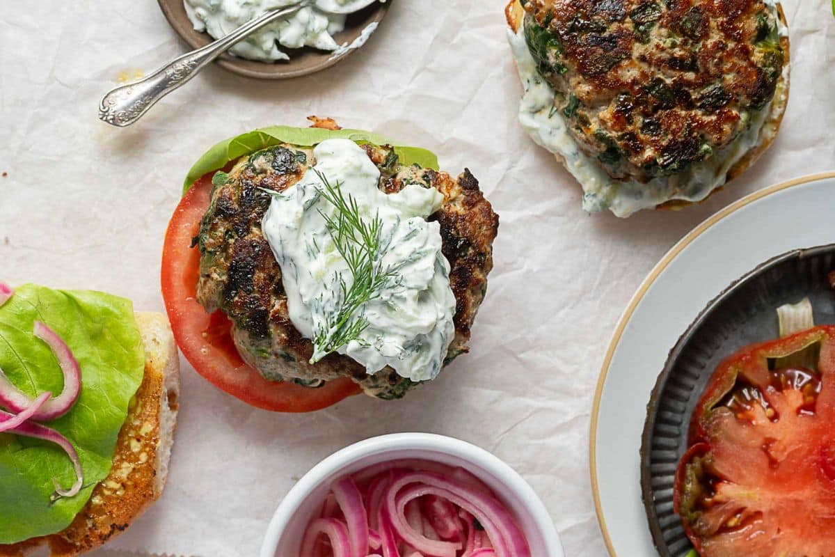 A turkey burger topped with tomato, lettuce, tzatziki sauce, and dill, surrounded by a bowl of pickled onions and sliced tomatoes on a white surface.