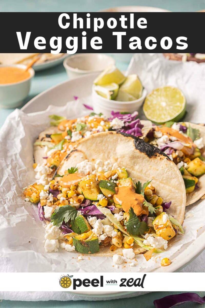 Two chipotle zucchini tacos with corn, cabbage, cheese, and cilantro are served on a white plate with lime wedges and a small bowl of sauce in the background.