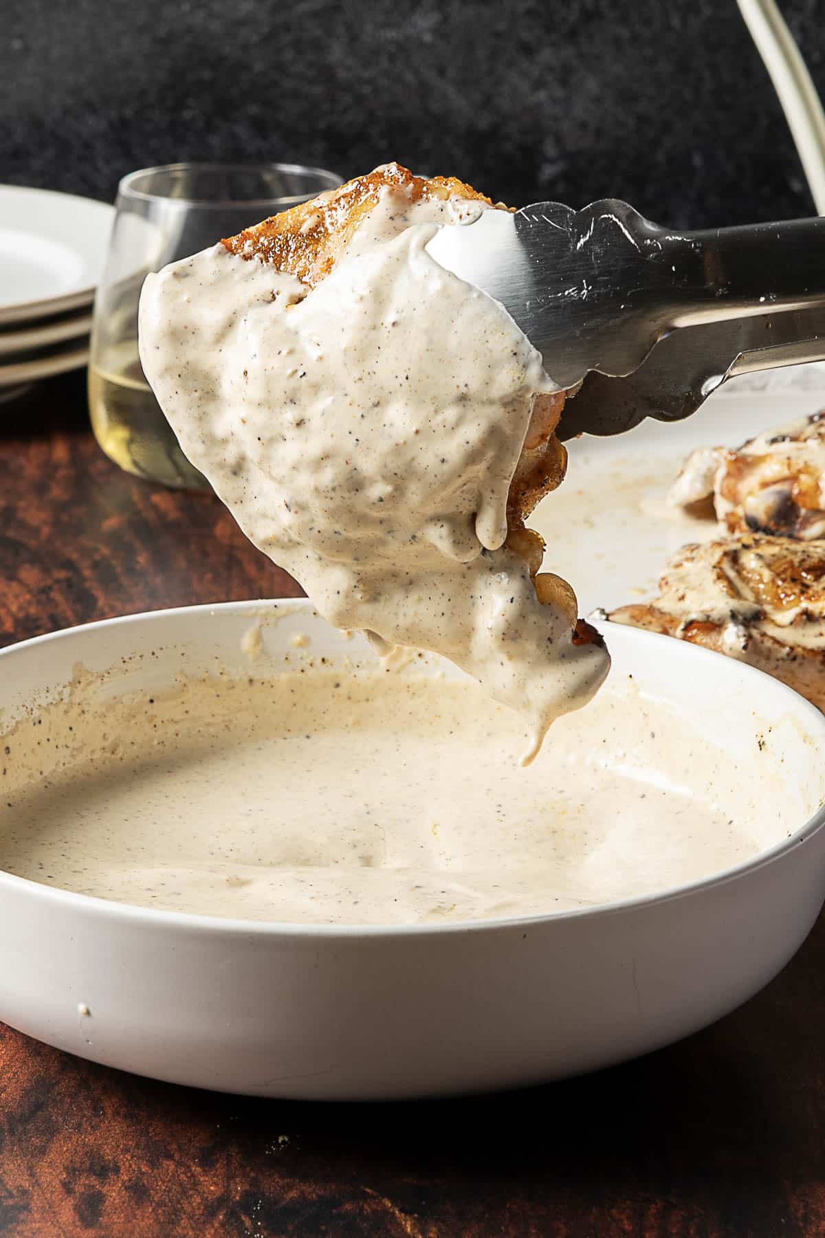 A piece of cooked chicken held by tongs being dipped into a bowl of creamy white sauce on a dark wooden table.