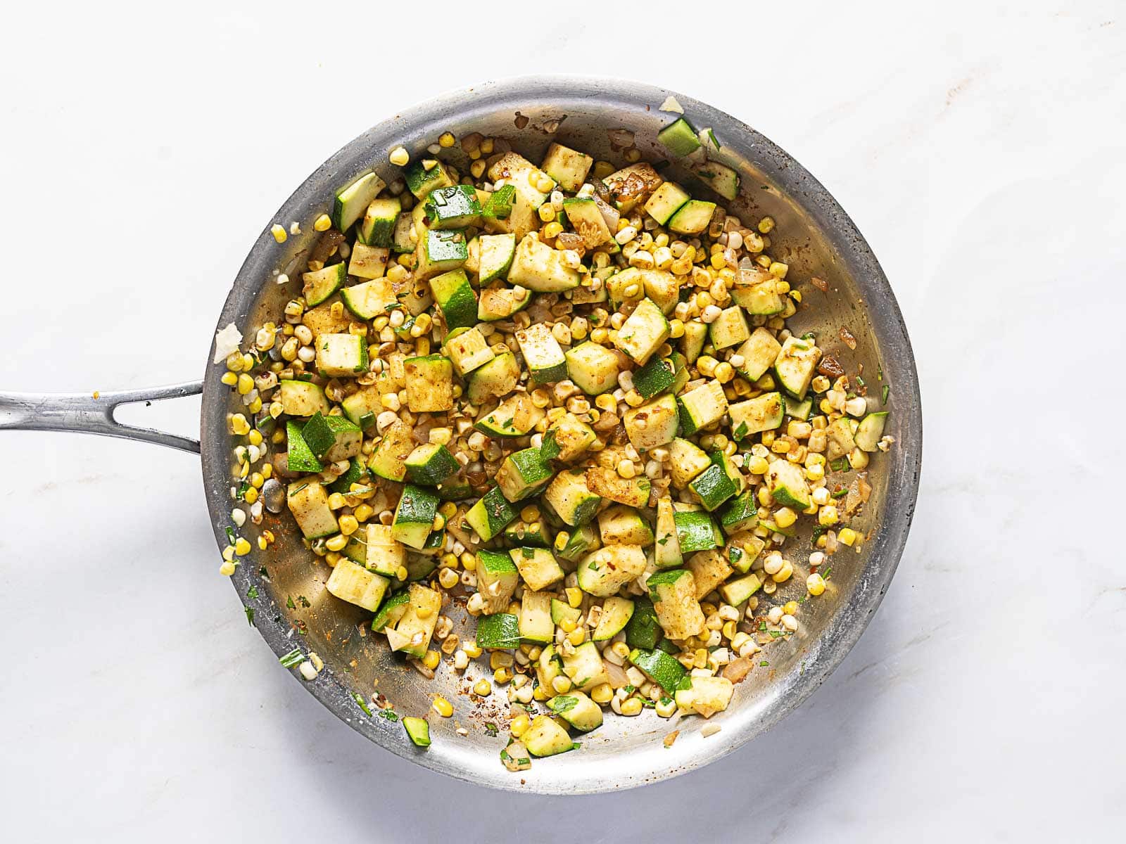 A skillet filled with sautéed diced zucchini and corn sits on a white surface.