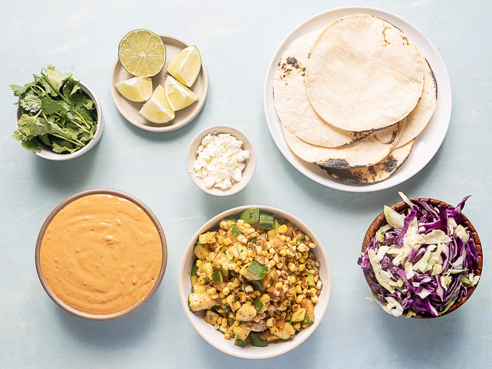 Overhead view of tortillas, cabbage slaw, corn and zucchini mix, chipotle sauce, cilantro, lime wedges, and crumbled cheese, all arranged in separate bowls and plates.