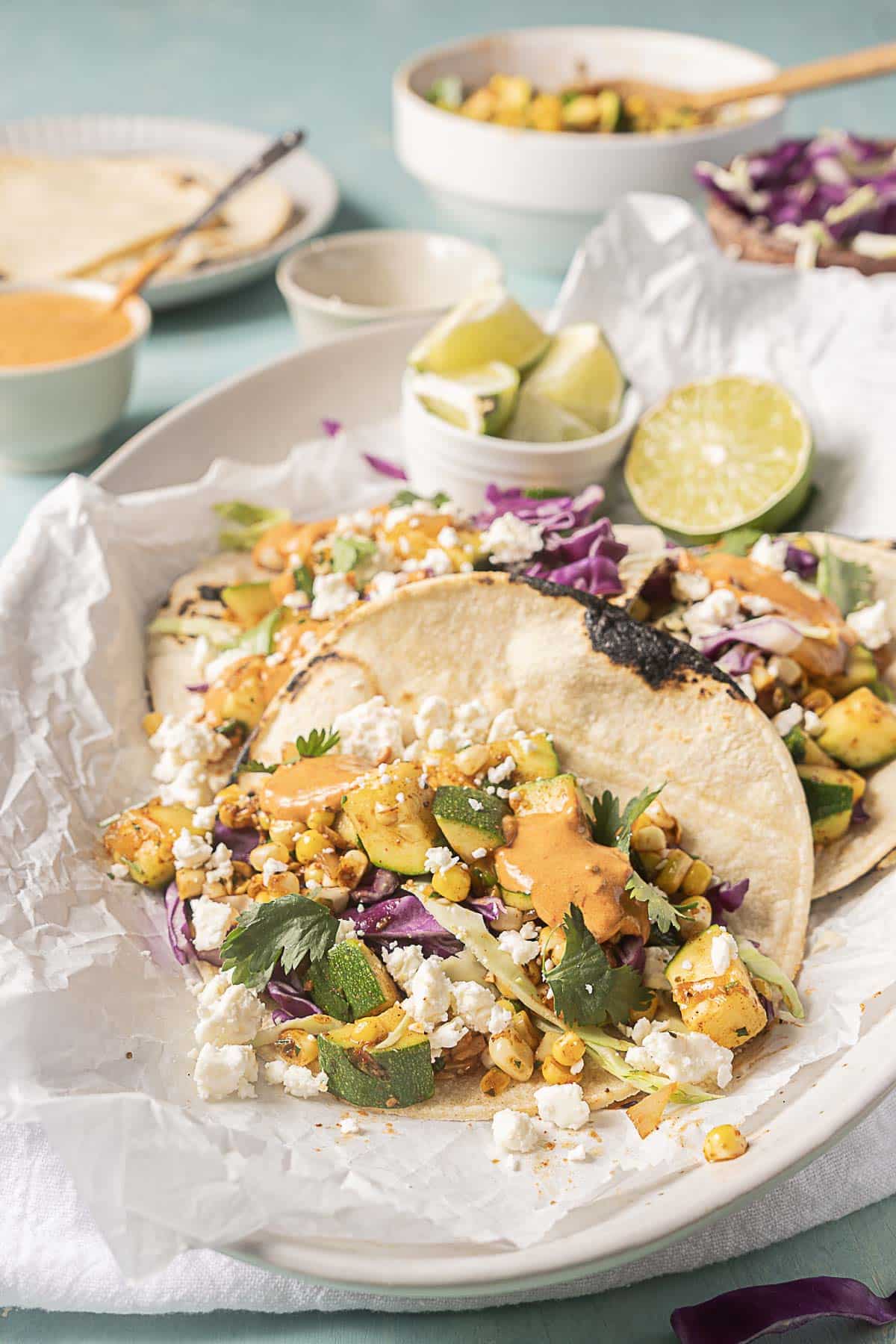Two vegetable tacos with grilled corn, zucchini, cabbage, cheese, and sauce on tortillas, served on a plate with lime wedges and extra sauce in small bowls.