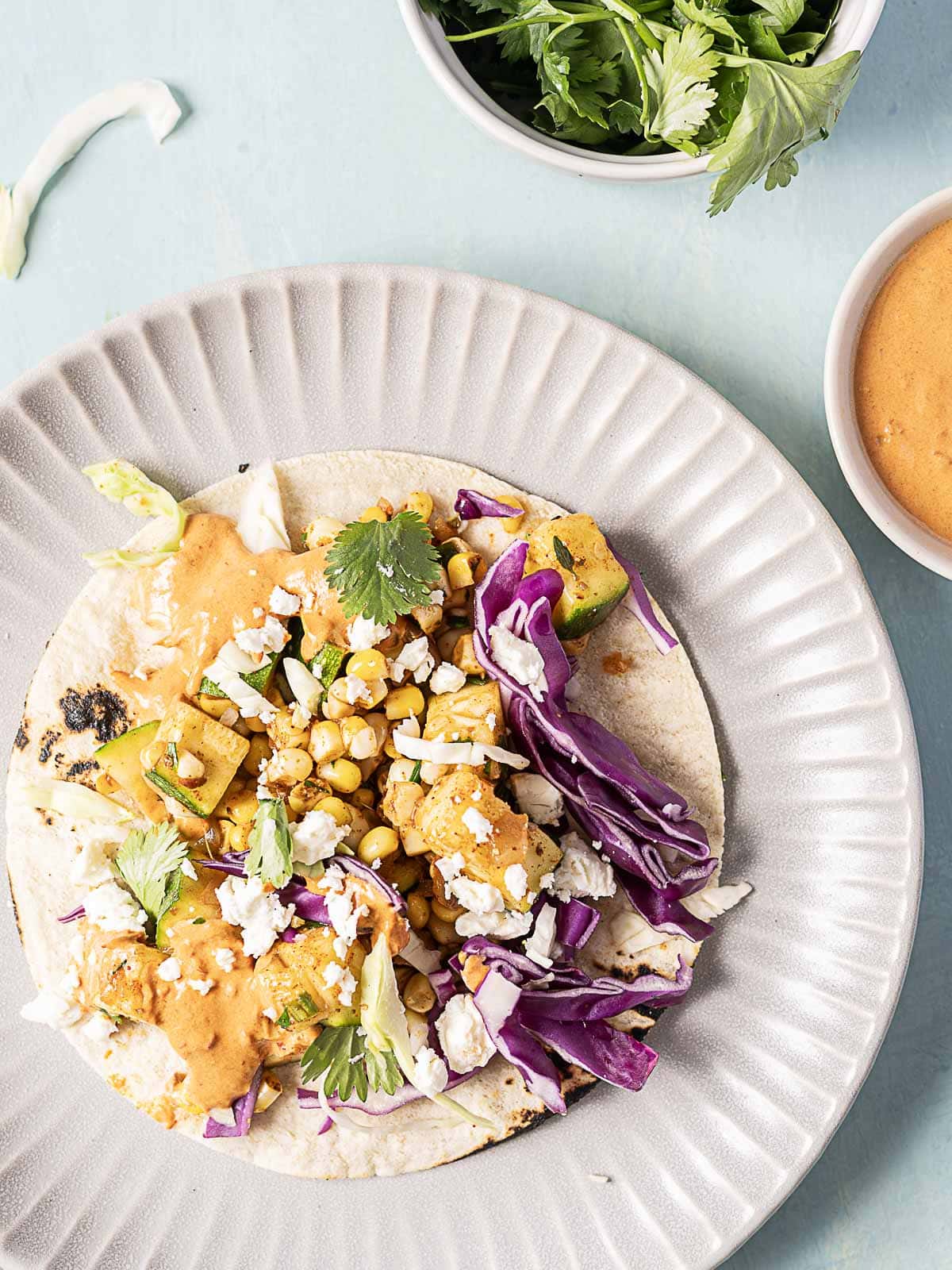 A taco with grilled vegetables, corn, cabbage, cheese, and cilantro sits on a plate. Small bowls of sauce and fresh cilantro are nearby.