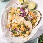 Three vegetable tacos with corn, cabbage, cilantro, cheese, and sauce are served on a platter with lime wedges on the side.