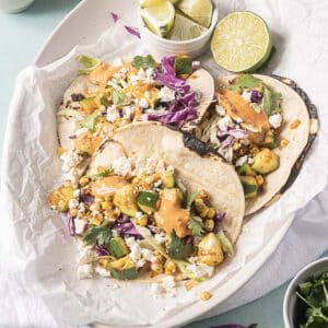 Three vegetable tacos with corn, cabbage, cilantro, cheese, and sauce are served on a platter with lime wedges on the side.
