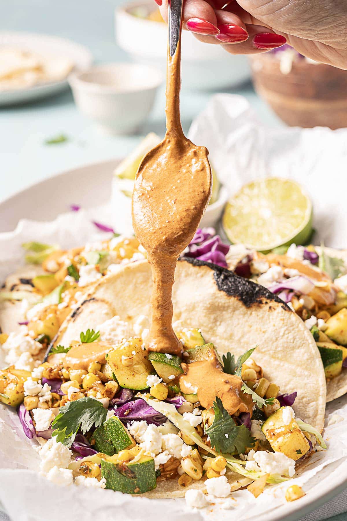 A hand drizzles creamy sauce from a spoon over vegetable tacos topped with corn, zucchini, feta, cabbage, cilantro, and lime on a white plate.