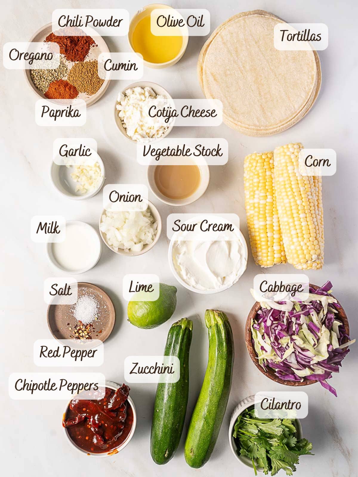 Assorted labeled ingredients for zucchini tacos, including spices, tortillas, cheese, sour cream, cabbage, corn, chipotle peppers, lime, onion, garlic, and cilantro.