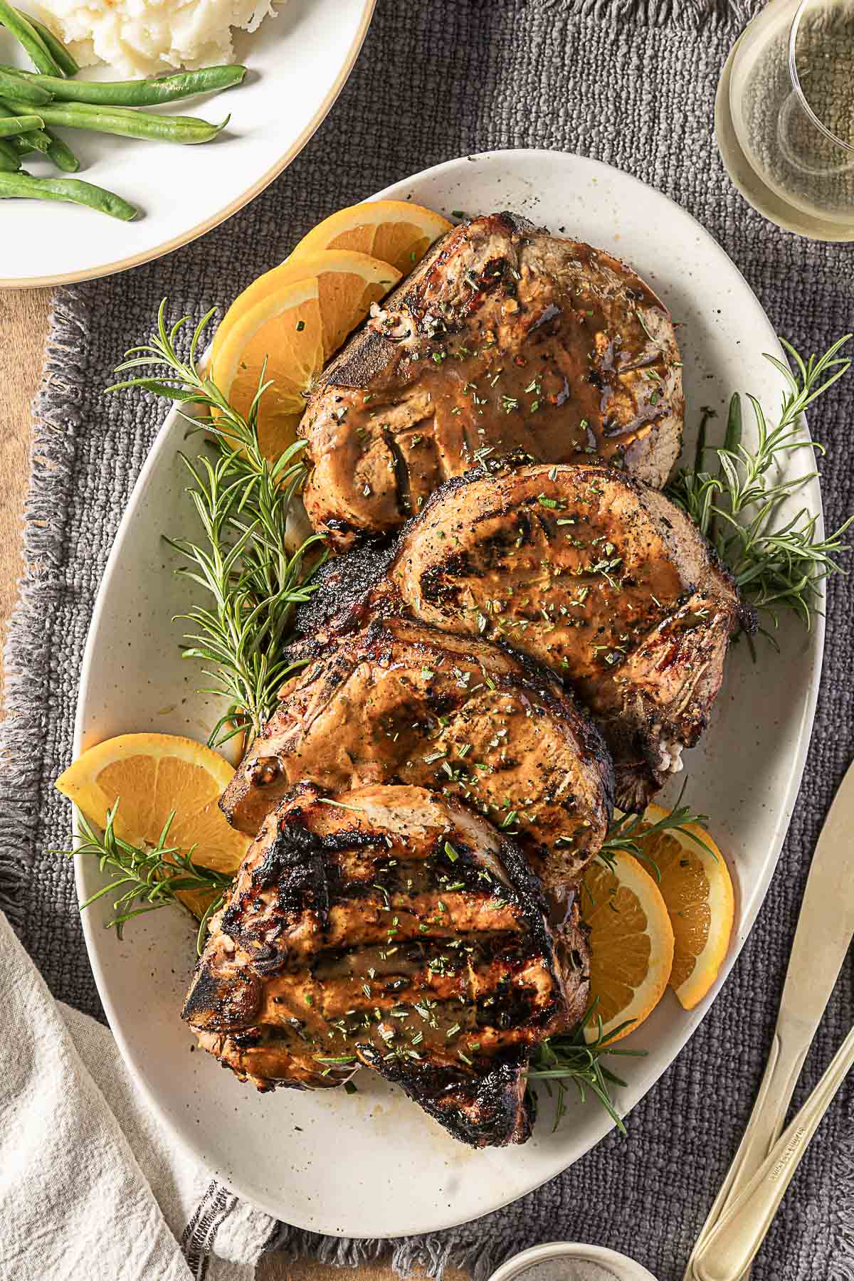 Grilled pork chops garnished with herbs and orange slices are arranged on a white oval platter, with rosemary sprigs placed around the meat.