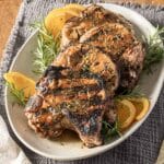 Three grilled pork chops garnished with orange slices and sprigs of rosemary on a white oval plate.