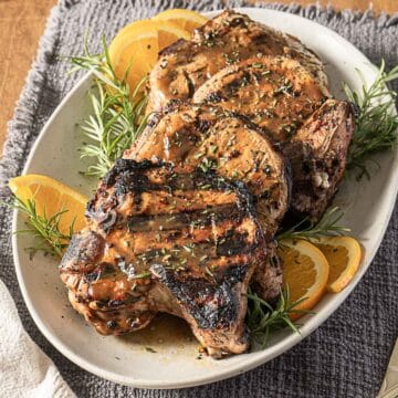 Three grilled pork chops garnished with orange slices and sprigs of rosemary on a white oval plate.