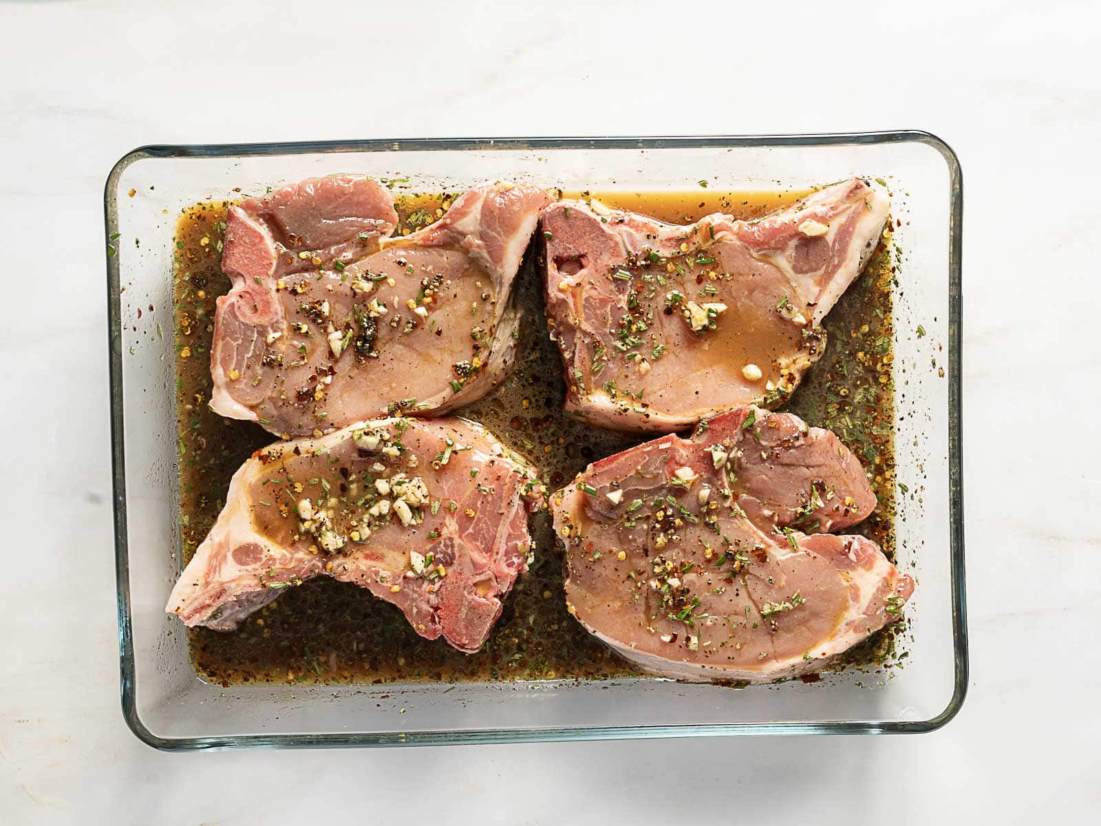 Four raw pork chops are marinating in a glass dish with a mixture of herbs, spices, garlic, and liquid marinade on a white surface.
