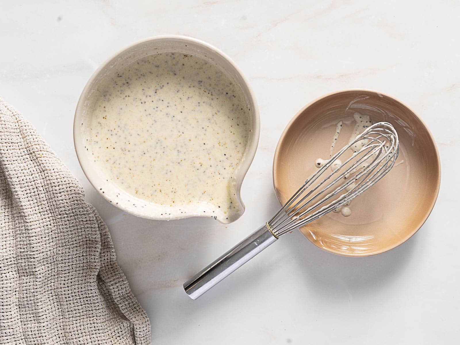 A bowl of creamy poppyseed dressing next to an empty bowl with a whisk, set on a light countertop with a textured cloth nearby.