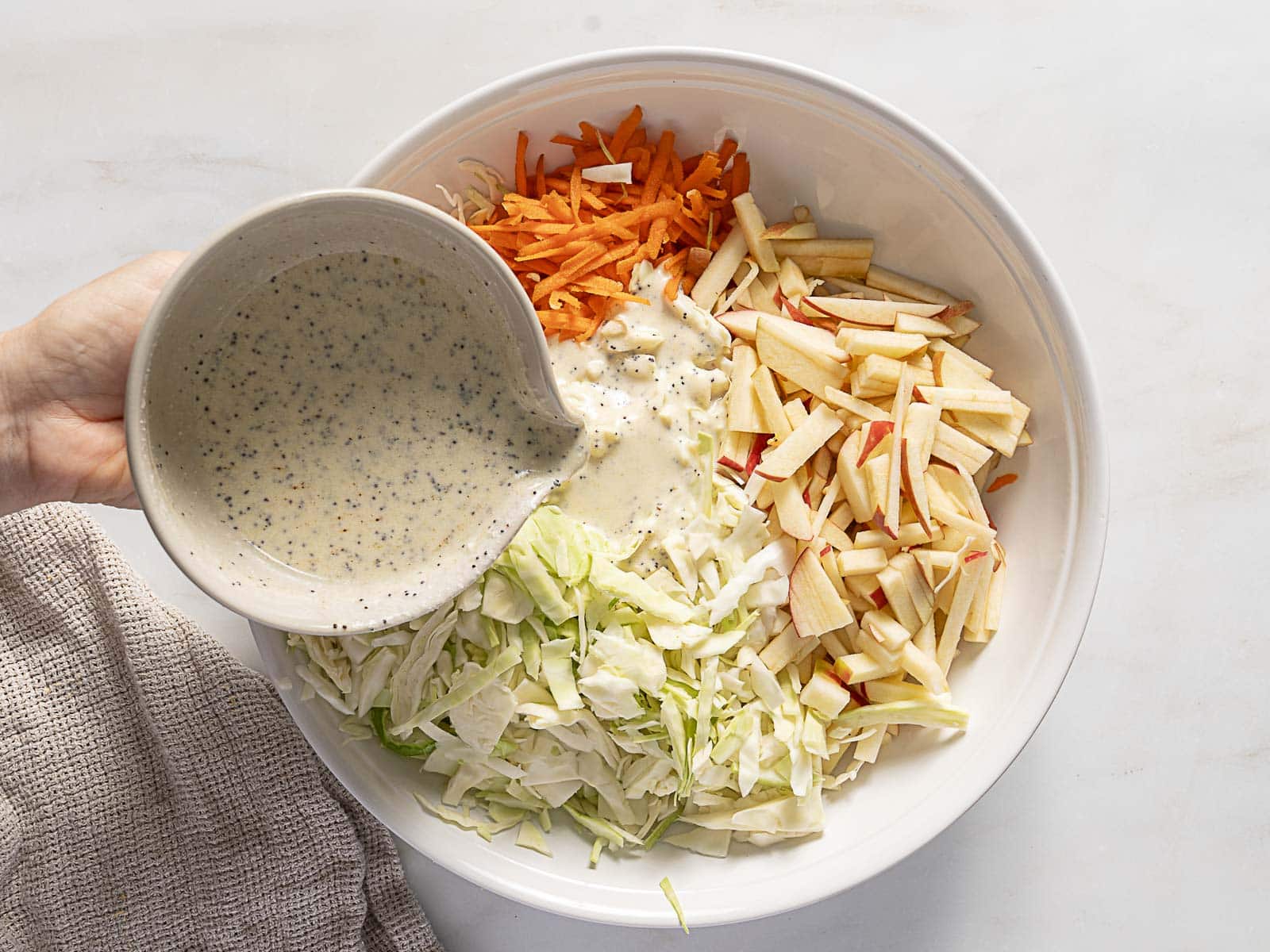 A hand pours creamy poppy seed dressing over a bowl of shredded cabbage, carrots, and apple slices.