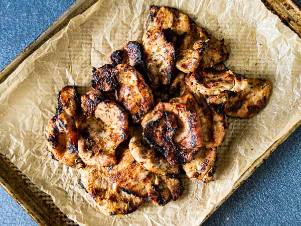 Grilled pieces of marinated pork are arranged on crumpled parchment paper atop a baking sheet.