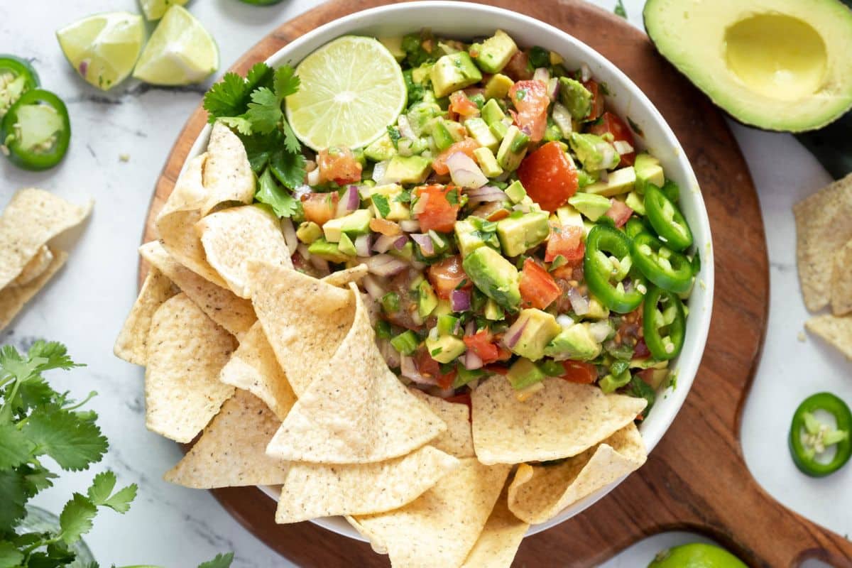 A bowl of avocado salsa with diced vegetables, ripe tomatoes, jalape&ntilde;o slices, and a lime wedge, surrounded by tortilla chips on a wooden serving board-a delicious addition to your must-try recipes.