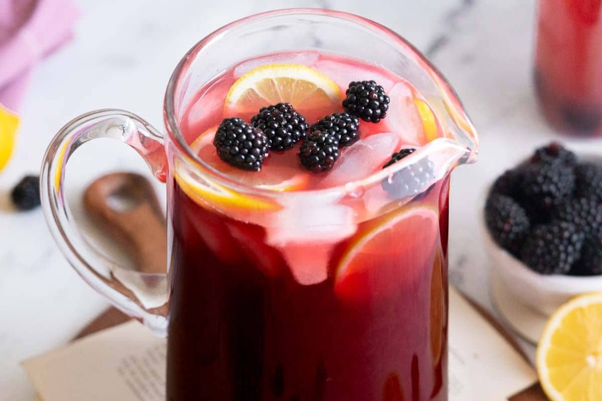 A glass pitcher filled with refreshing red iced tea, blackberries, and lemon slices. Perfect for summer drinks, with a bowl of blackberries and a lemon visible in the background.